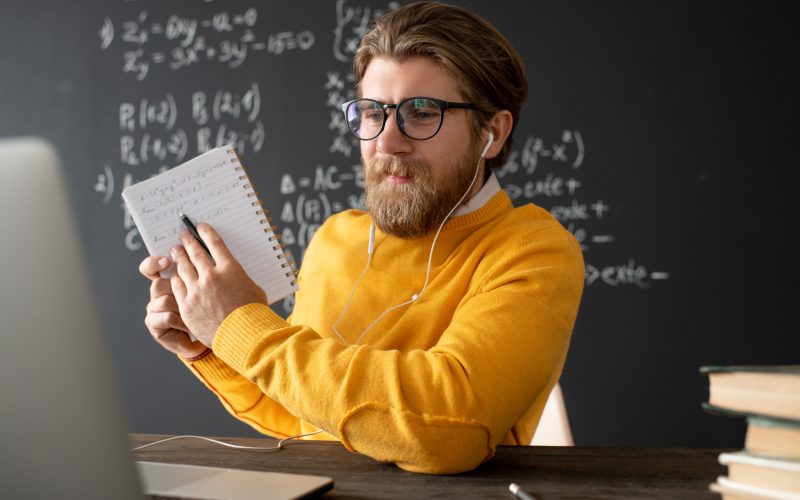 Young confident teacher in eyeglasses and pullover pointing at his notes in copybook while explaining them to online audience in front of laptop